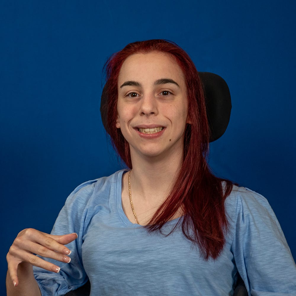 A light skinned woman with dark hair and a light blue shirt in a wheelchair smiling at the camera.