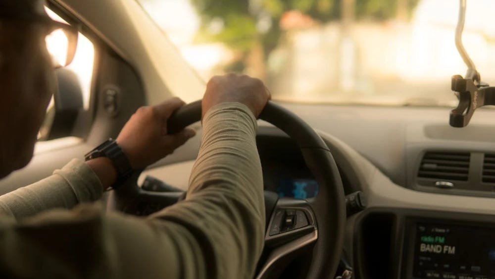 Fotografia de uma pessoa dirigindo um carro. Na imagem é possível ver apenas parte do painél do veículo. A pessoa está com as duas mãos no volante, olhando para frente.