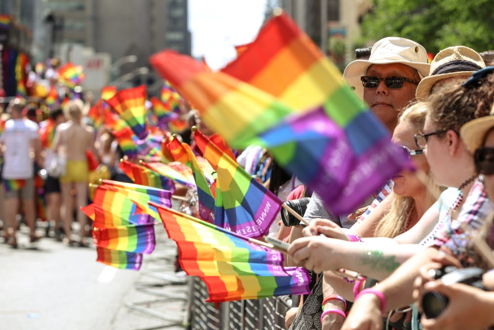 photo d’un trottoir bondé bon-enfant familial blanc tenant tous et toutes le même drapeau LGBT fait en série, derrière une barrière en métal qui les sépare de l’avenue vide où on aperçoit au fond un défilé flou. Le tableau ressemble à celui du défilé militaire d’une fête nationale.