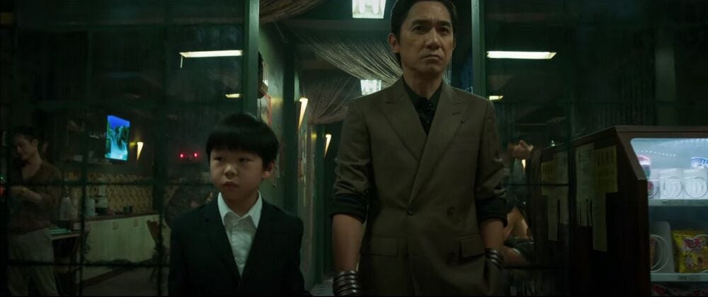 Wenwu, with his sleeves rolled up and his ten rings visible on both arms, holds the hand of seven-year-old Shang-Chi in a hallway.