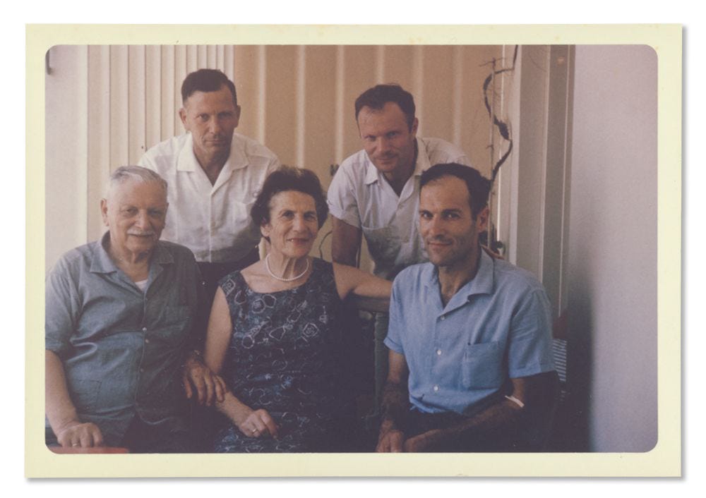In a color portrait of a family, an older man and woman sit in the front row to the left of a young man wearin a blue short-sleeve shirt. The older man is wearing a blue button-down shirt and the woman is wearing a blue-pattered sleeveless dress and a white pearl necklace. Behind them, leaning on the chairs to be in the frame, are two other young men wearing white, short-sleeve shirts.