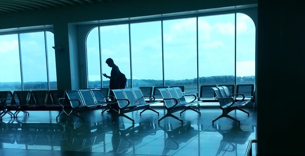 Foto de uma sala de espera de aeroporto praticamente vazia. Na sala há apenas apenas uma pessoa. É um homem, que está em pé, olhando cabisbaixo para o seu celular.