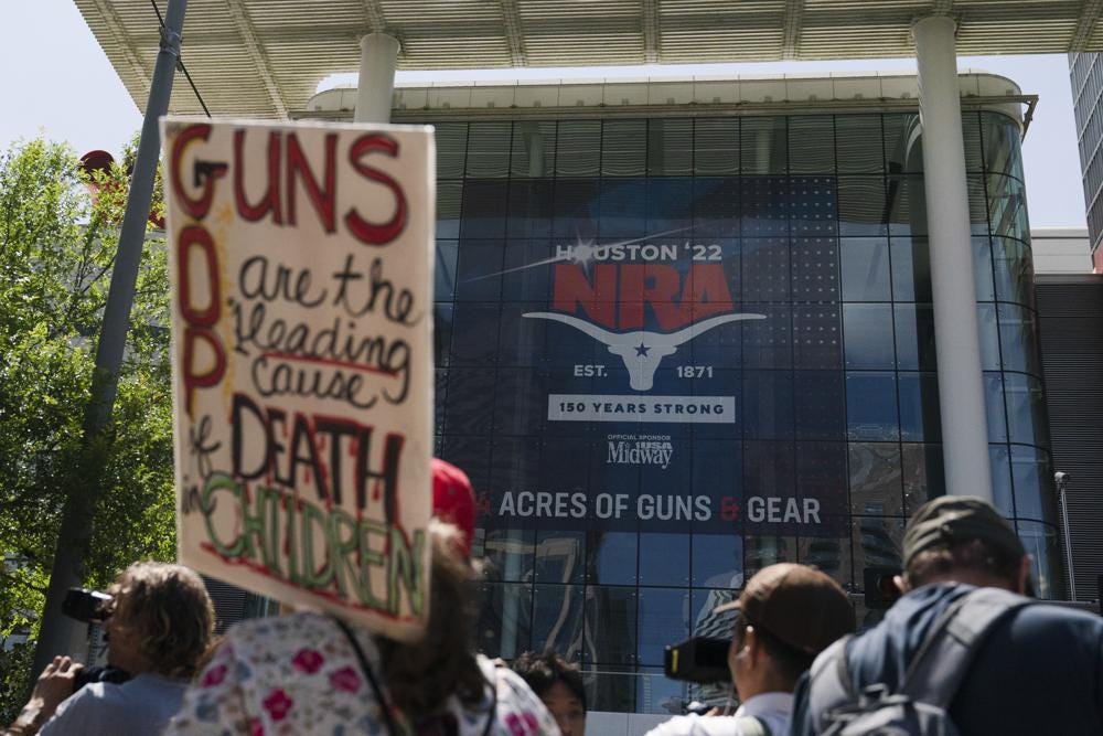 A protestor holds a sign with “GUNS” and “GOP” written on it, with the words sharing the “G.” It reads, “GUNS/GOP are the leading cause of DEATH in CHILDREN.” They are standing in front of a convention center with a sign for the 2022 NRA Annual Meeting.