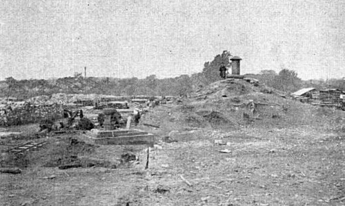 Grainy old photo of bare ground with a small square monument on the left and a mound on the right. A man stands on the top of the mound beside a taller stone monument.