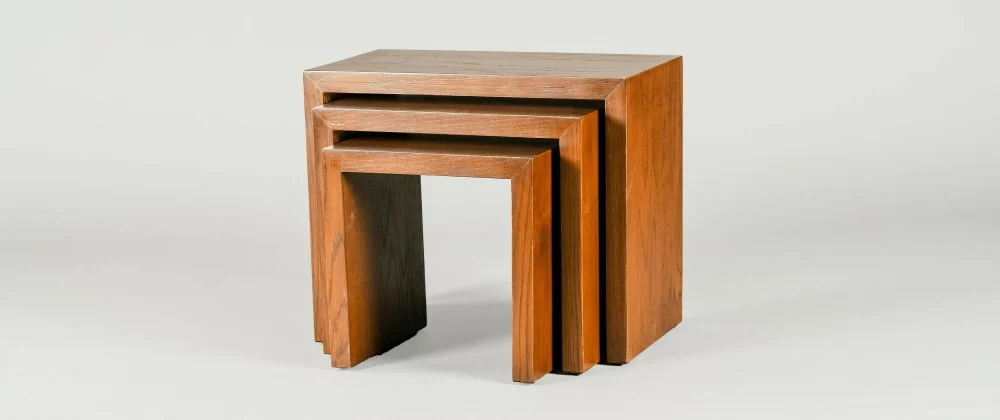 A set of three nested wooden tables of descending size, displayed in a staggered arrangement to show how they fit together. The tables have a smooth finish with a natural wood grain, and they are photographed against a neutral background, highlighting their simple, clean lines and functional design.