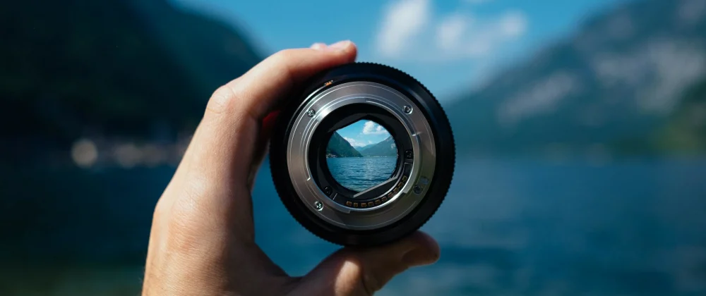A hand holding a camera lens against a scenic landscape, with the lens framing and focusing on a mountain peak and a body of water. This perspective creates an interesting visual effect, where the landscape appears sharp within the lens and blurred outside it, emphasizing the concept of focus and the art of photography.