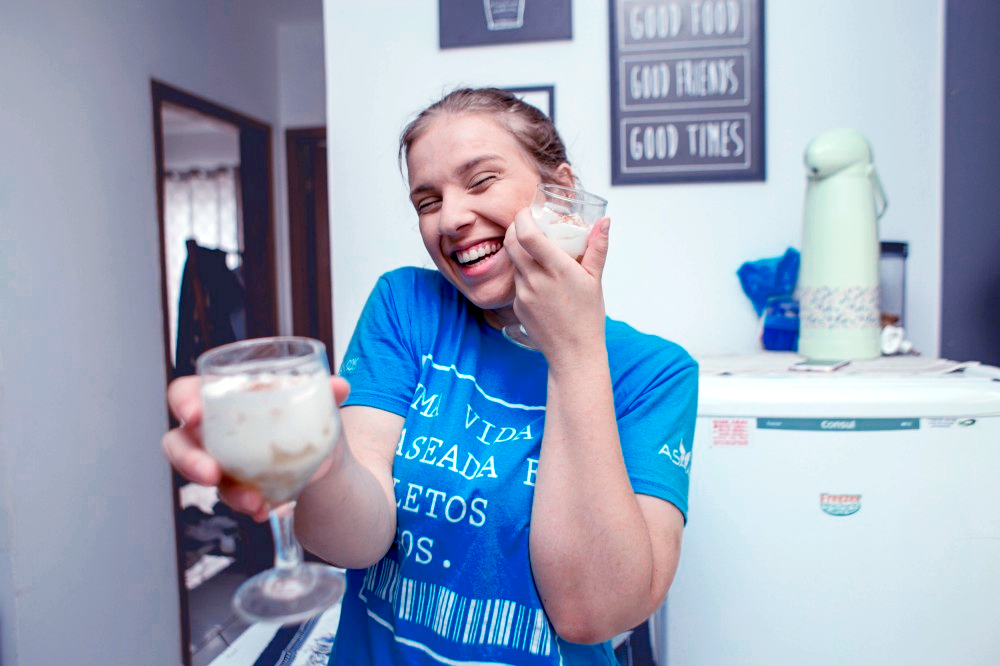 Carolina Oleskovicz, staff na equipe de Empreendedores do Asaas, sorri para a câmera, com os olhos entreabertos, enquanto segura duas taças contendo a sobremesa feita de abacaxi com chantilly e canela. Carolina é loira e veste uma camiseta na cor azul claro, com a frase “Uma vida baseada em boletos pagos” estampada.