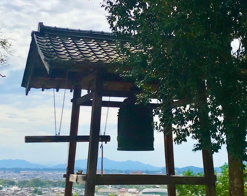 A large cylindrical bell with round wooden knocker hung horizontally beside it are silhouetted against a cloudy sky. The bell is housed under a tile roof. There are leafy trees on the side and mountains in the distance.
