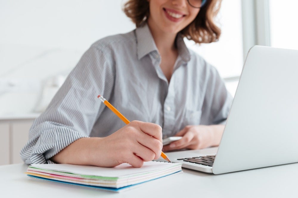 young copywriter woman in striped shirt taking notes while sitting at table in light apartment