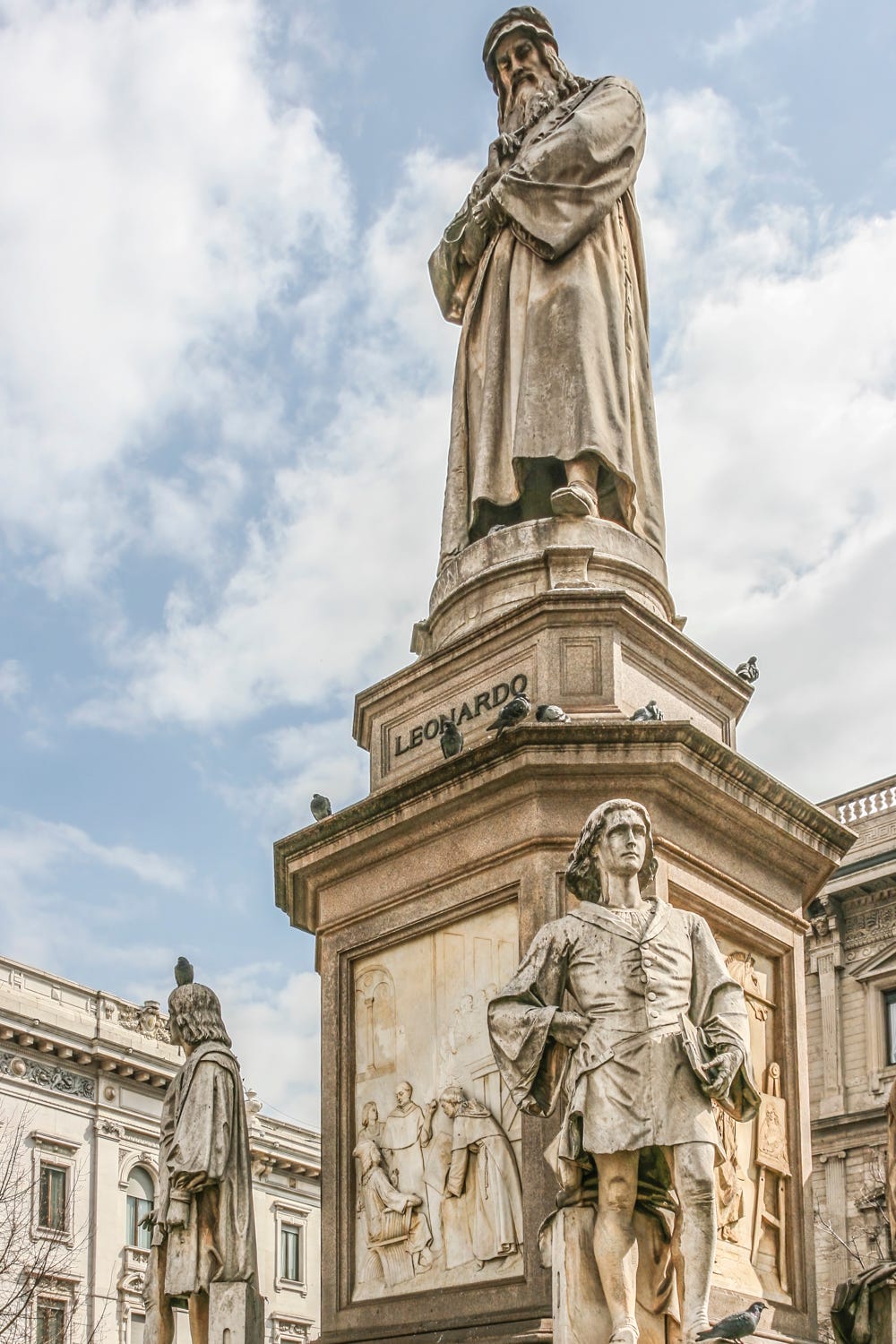 The Leonardo monument in Milan’s Piazza alla Scala, showing Leonardo on top and two of his four students below him. There are bas-reliefs between the students that demonstrate the diverse interests of Leonardo.