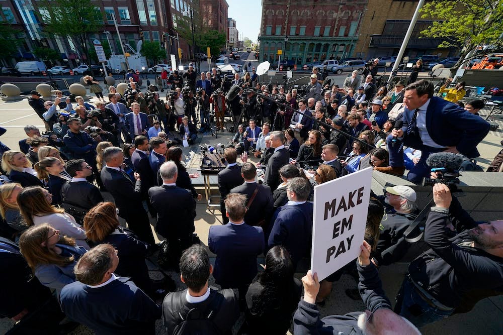 A large group of people and press in a scrum, with someone holding a sign saying “Make em pay”