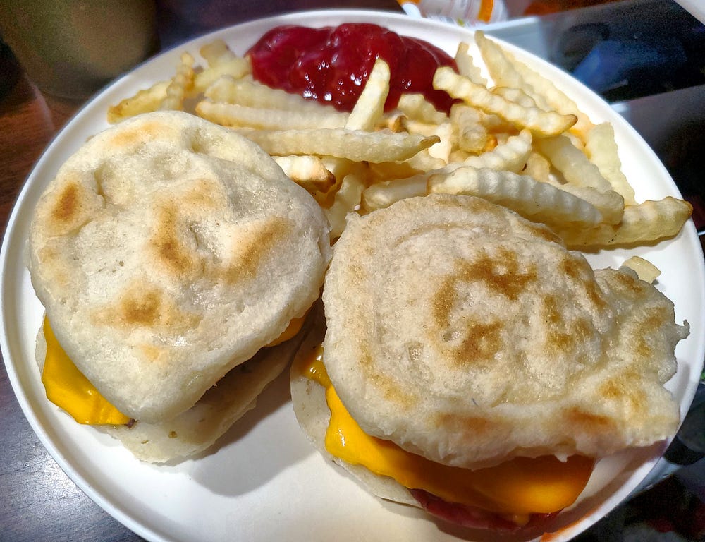 Two cheese burgers with split English muffins as the buns sit on a plate with side of crinkle cut french fries and ketchup.