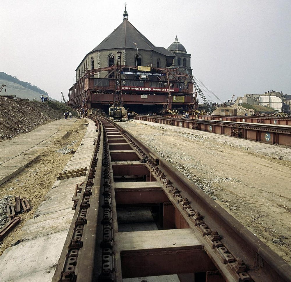 Color photograph showing a large building, enclosed in a metal structure, being moved on rails