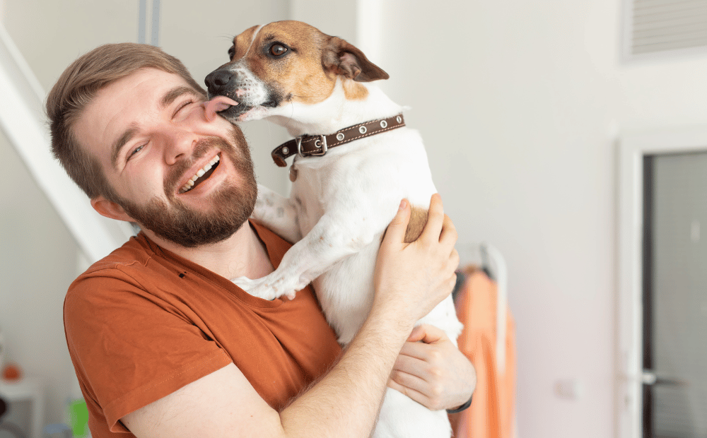 A smiling man holding a happy dog that is licking his face, both enjoying the benefits of improved heart health.