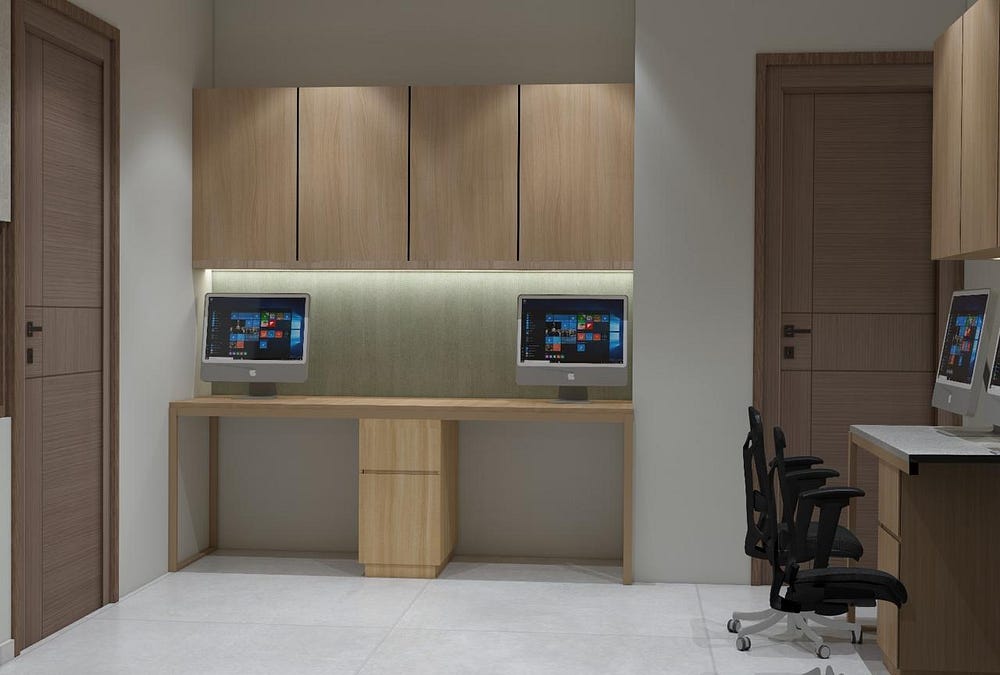 This office interior showcases a contemporary dual workstation setup, designed for efficiency and collaboration. The space features clean wooden cabinetry with concealed storage, ambient under-cabinet lighting, and neatly arranged desktop computers. Ergonomic black mesh chairs complement the minimalist aesthetic, ensuring comfort during long work hours. With its balance of style and practicality, this modern office design fosters productivity in a clutter-free environment.