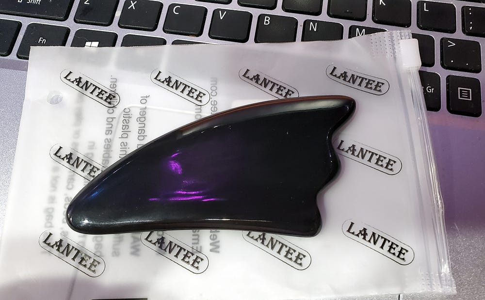 A triangular shaped piece of smooth dark material with slightly curved edges and a wavy end sits on a plastic envelop with the word Lantee printed in rows. The items are sitting on a laptop keyboard.