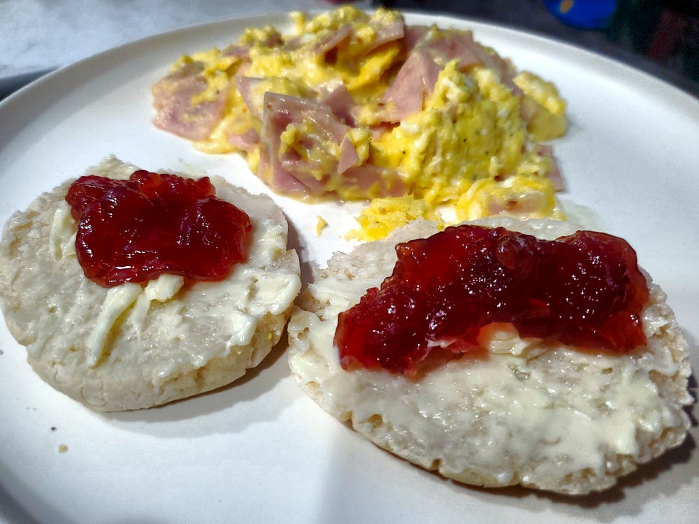 Two halves of a split English muffin topped with butter and strawberry jam sit on white dinner plate. Behind the muffins is a helping of scrambled eggs with cheese and ham.