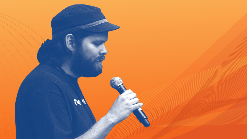 Grey-tone photo of Kin Lane wearing a hat and talking into a mic against an orange background.