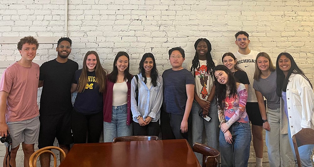 Diverse group of 12 young men and women from the University of Michigan posed in front of a white brick wall.