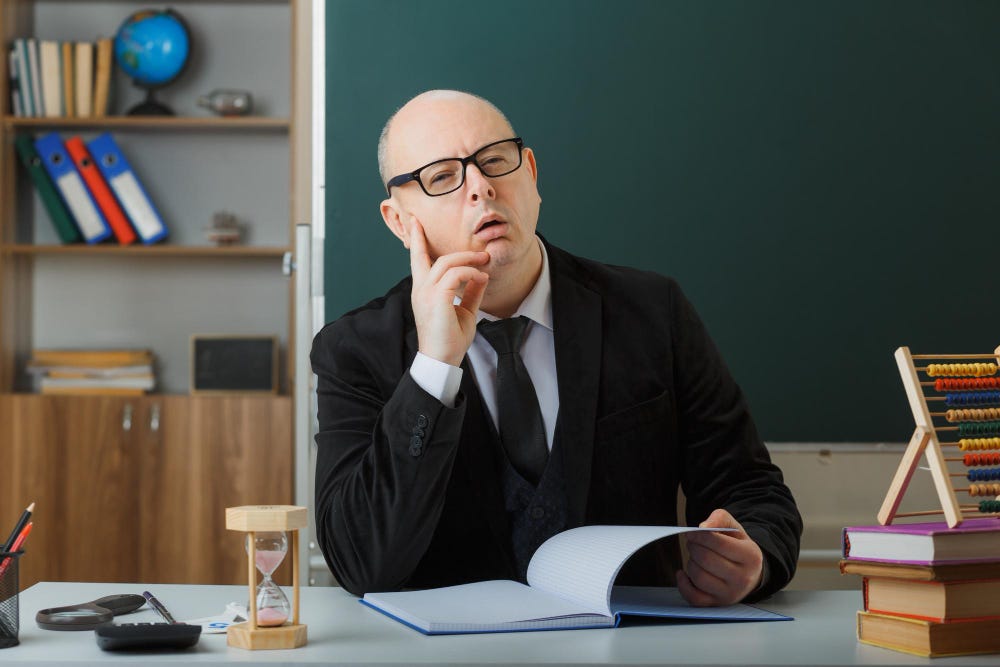 A pale man seated at a teacher’s desk looks confused.