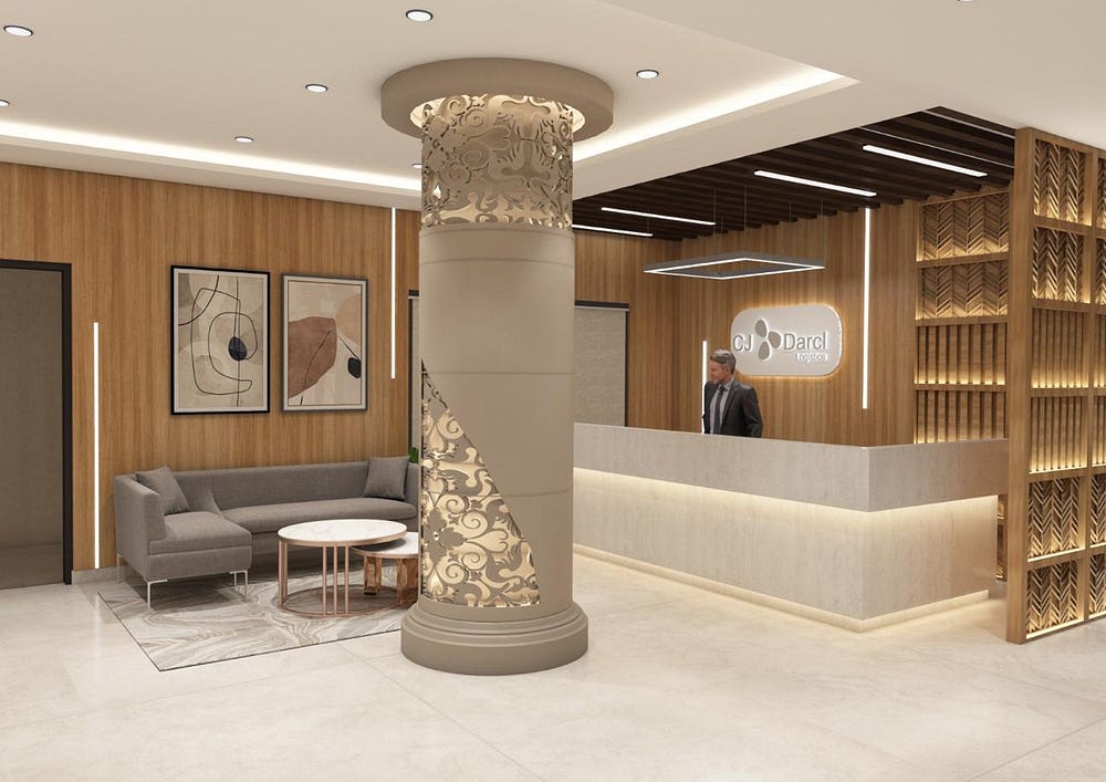 A refined hotel reception area featuring a modern front desk with warm wood accents and soft underlighting. A striking decorative pillar with intricate golden cut-out patterns serves as a centerpiece. To the side, a cozy grey sectional sofa with marble-top nesting tables offers a stylish waiting space, enhanced by abstract wall art and a patterned rug. The overall ambiance combines sophistication with comfort, creating a welcoming first impression for hotel guests.