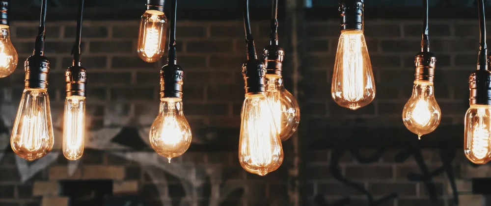 A row of vintage-style Edison light bulbs hanging from black cables against a dark brick wall background. The bulbs emit a warm, glowing light that creates a cozy and inviting atmosphere, while highlighting the texture of the wall behind them. The image conveys a sense of contemporary interior design with a nod to retro aesthetics.