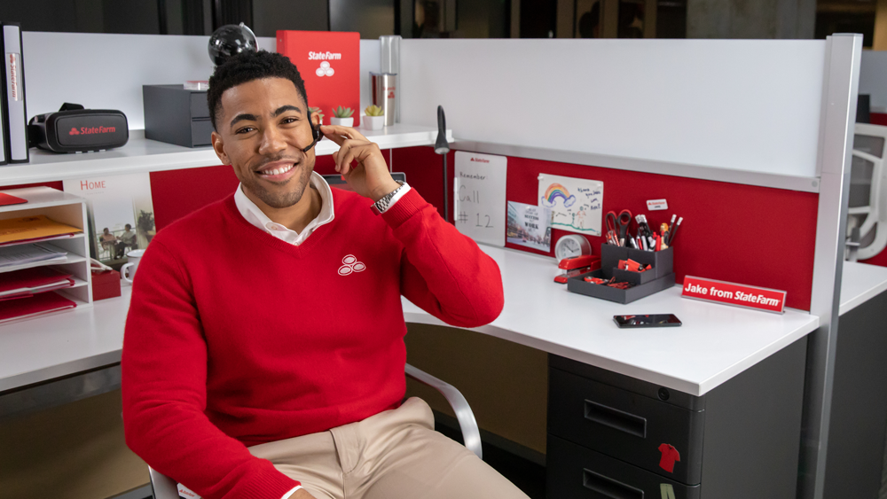 Jake from state farm in a red sweater smiling at his desk.