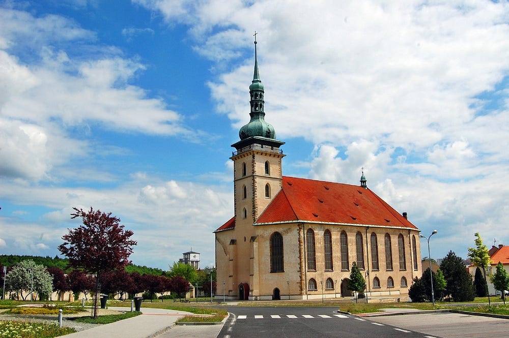 Modern photograph showing a Gothic church with a red roof and green onion domes, recently restored. The building is made of lightly colored bricks and features high windows