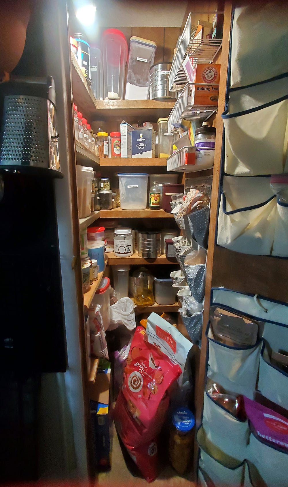 A narrow pantry with a light in the upper left. There are shelves in the back and on the left side. On the right on the wall is a white wire shelf unit and hanging pocket shoe storage. Similar shoe hanging storage is attached to the pantry door. The shelves and hanging pockets are filled with pantry goods. Bags of dry cat food, boxes of garbage bags and other items are on the floor.