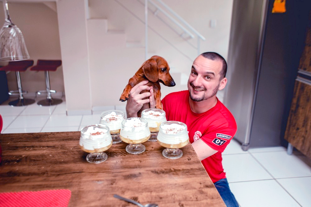 Giorgio Tamanini, teclíder de treinamentos no Asaas, sorri para a câmera enquanto segura um cachorro. Os dois estão perto de uma mesa, sobre a qual estão 5 taças contendo uma sobremesa de abacaxi com chantilly e canela. Todas as taças estão bem cheias. Giorgio possui o cabelo curto e veste uma camiseta vermelha.