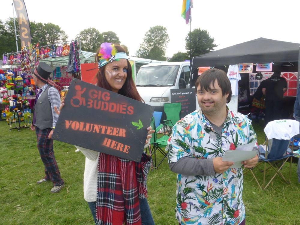 Man and woman at a festival holding a sign saying ‘Gig Buddies — volunteer here’