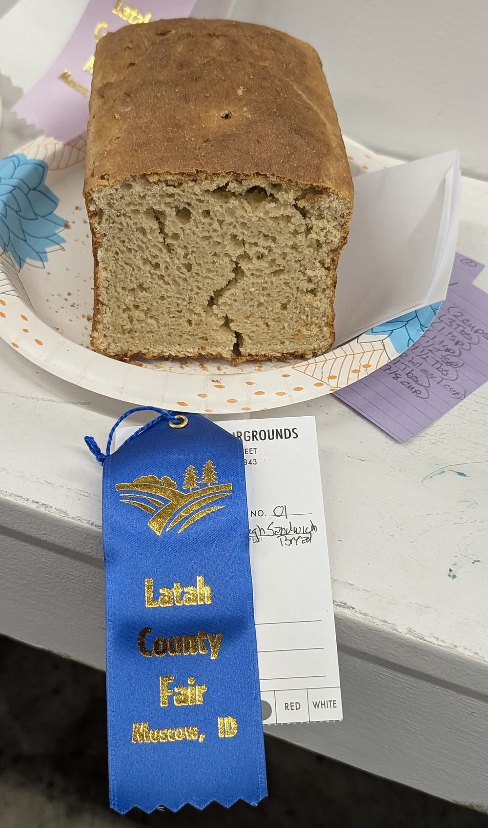 A loaf of bread sits on a white paper plate, adorned with a blue ribbon. The ribbon indicates a prize at the Latah County Fair.