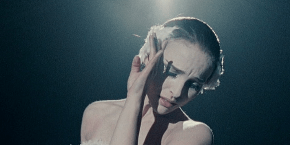 An image shows the actress with white feathers on either side of her head. She has a sad, strained emotion on her face and shields her face from a light that is pouring in from the top.