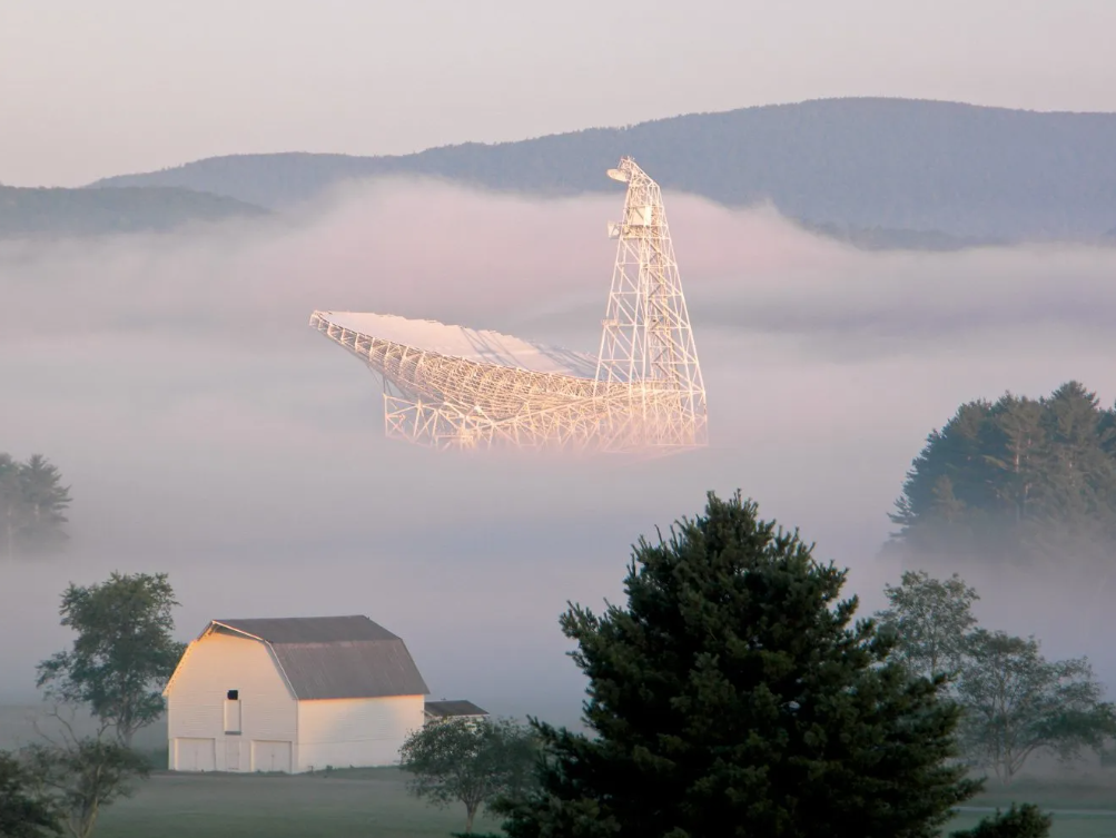 Photograph of the Green Bank Telescope nestled in morning mist. In the foreground is a barn and several trees; behind lie the hills.