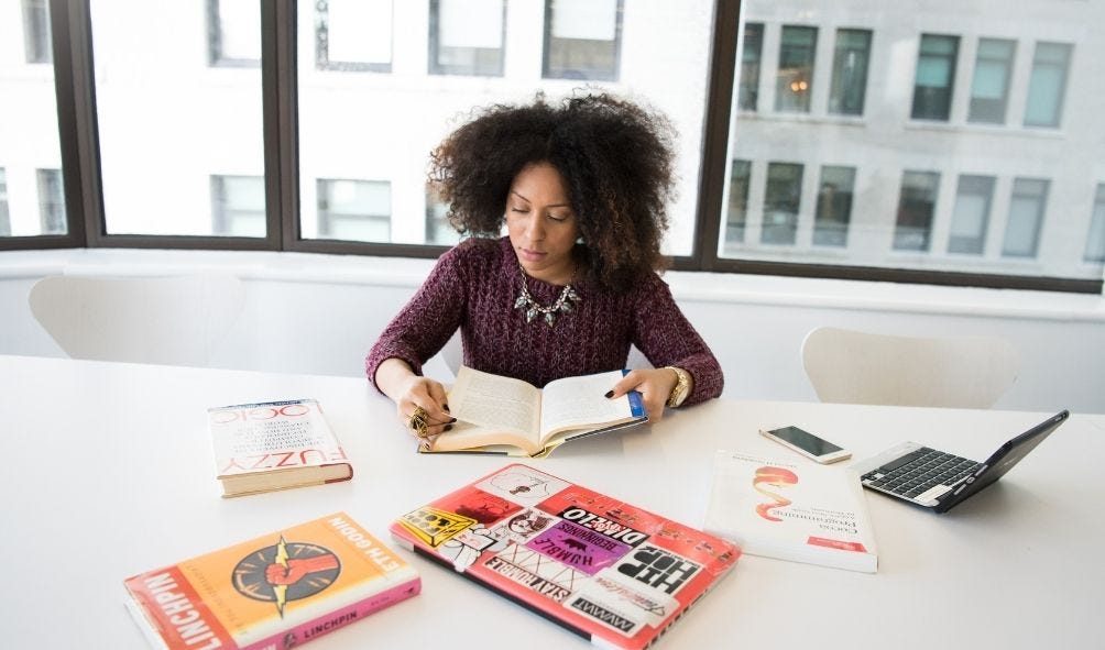 Imagem de uma mulher negra, sentada em uma cadeira branca, lendo um livro sob uma mesa de reunião com outros livros e anotações nos tons de amarelo, laranja, vermelho e rosa, celular e notebook.