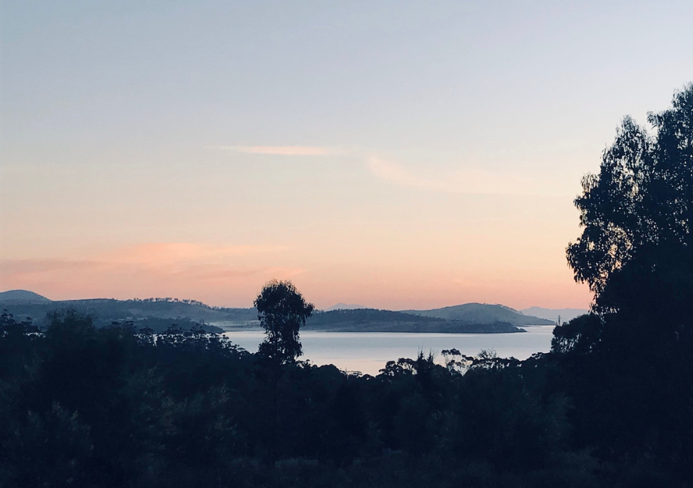 View of a bay in Tasmania at Dawn with a pinkish orange sky and silhouetted trees. There are views of the headland opposite but they are very dark.