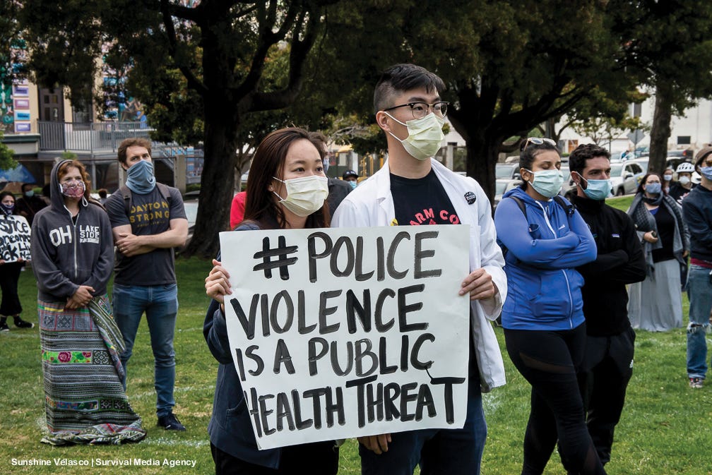 An Asian couple wears masks & scrubs, holding a sign: #Police Violence is a Public Health Threat.” Credit: Sunshine Velasco.