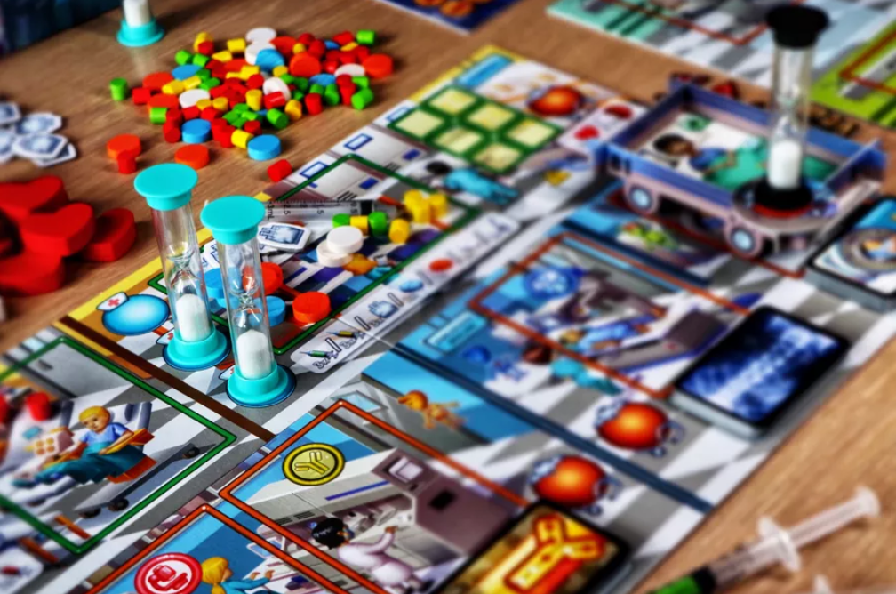 The game Rush M.D. Sand timers are positioned around a board with many small, colored cubes, fake syringes, and wooden hearts. One sand timer is on a cardboard hospital bed.