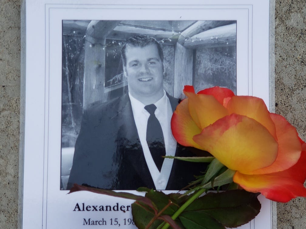 Black and white photo of a memorial brochure, showing Alex,  who died by suicide in 2012. With a yellow rose from the author’s garden…
