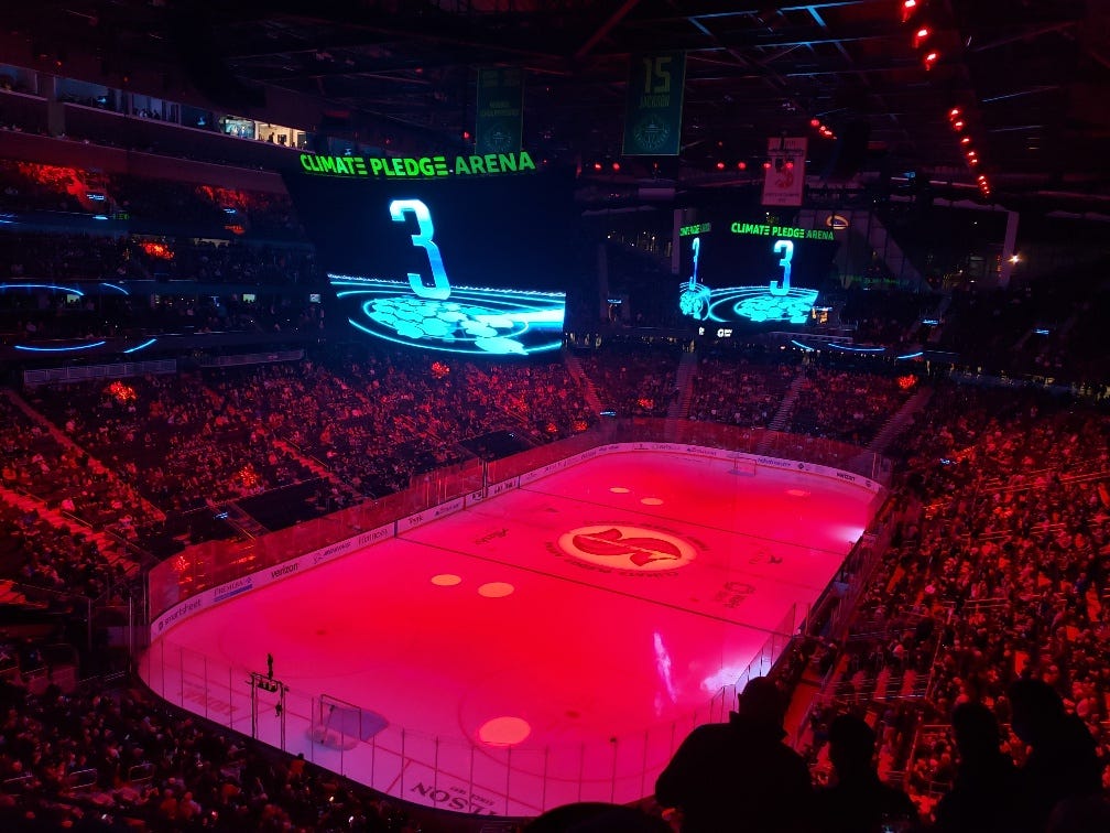 Inside the Climate Pledge Arena, dual state-of-the-art digital scoreboards float in the air above an ice rink glowing bright red, amongst a packed house.