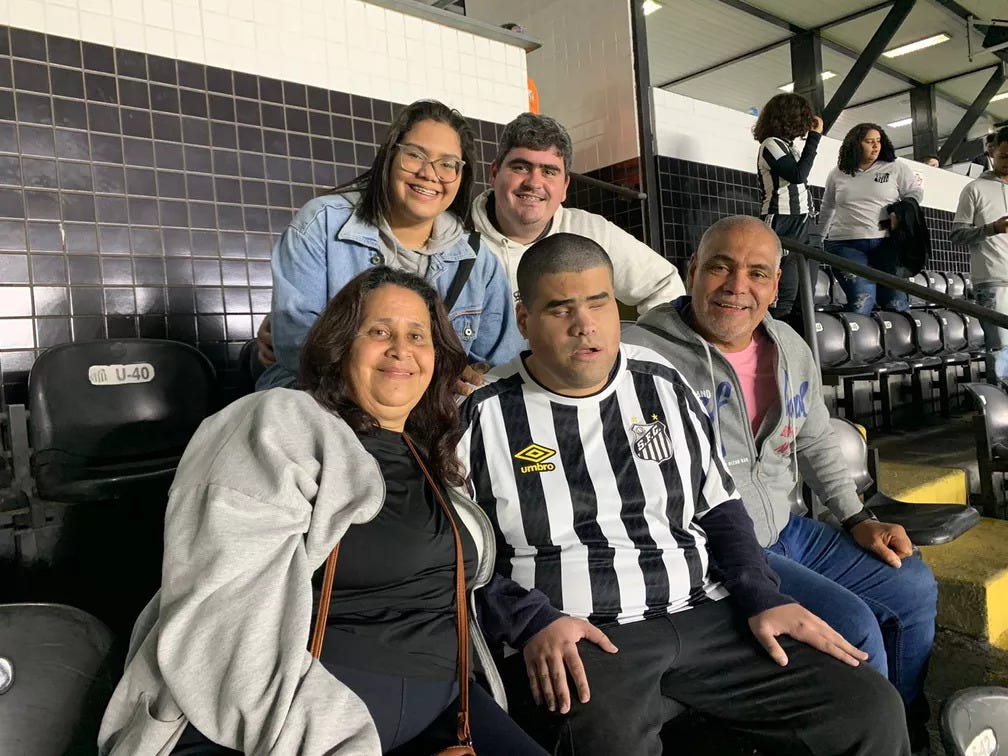 Foto do torcedor santista Matheus e sua família nas arquibancadas da Vila Belmiro, estádio do Santos. Na primeira fileira, Matheus está sentado entre seu pai e sua mãe, com outros dois familiares do torcedor na fileira de trás.