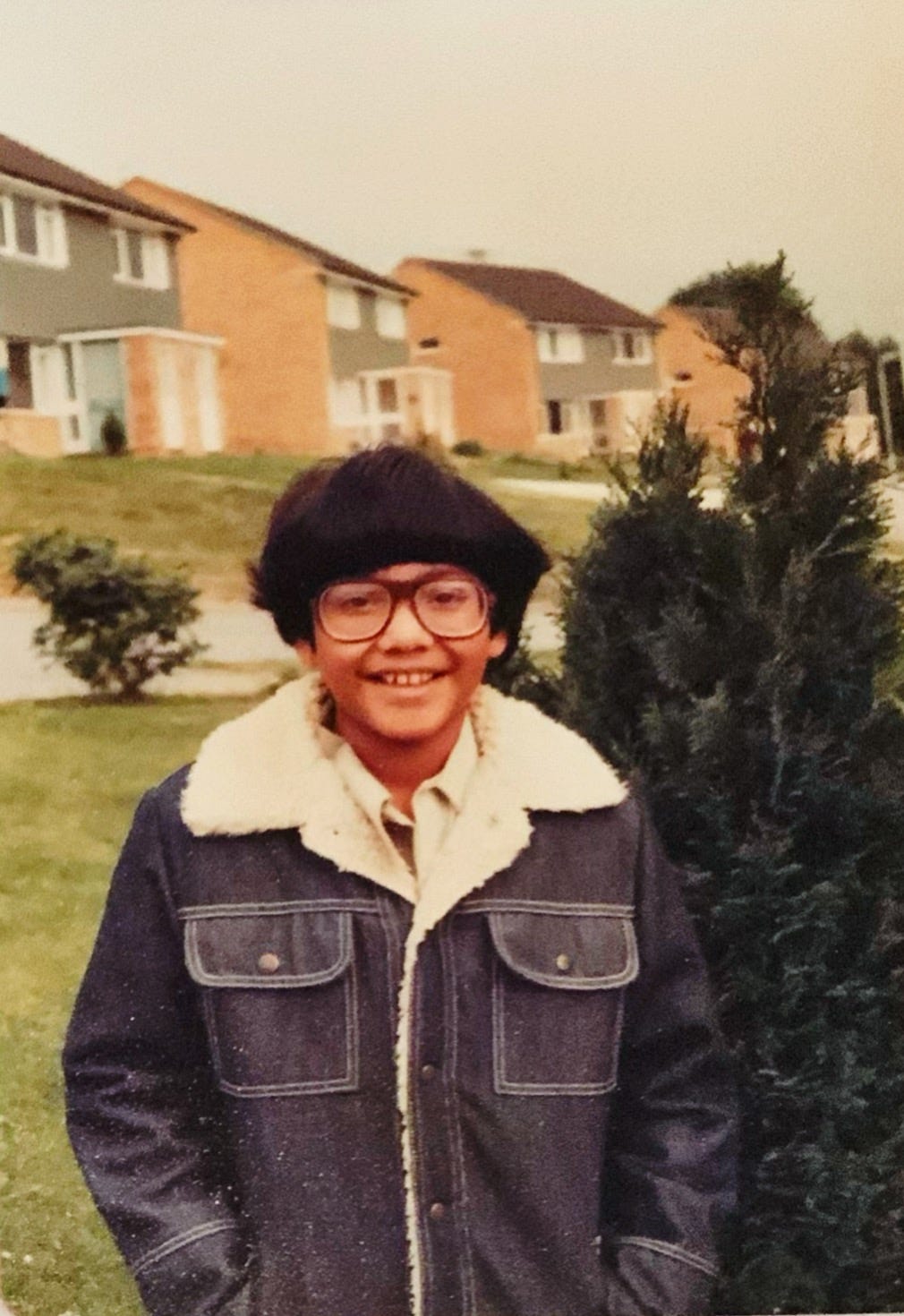 An early color photograph shows young brown skinned boy in thick 1970s glasses, and a bowl haircut, smiling at the camera. He wears a dark blue denim jacket with a fleece lining. He is standing outside in front of a row of houses.