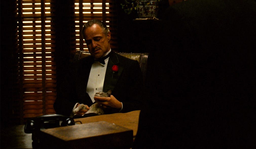 Temos uma mesa com um telefone e atrás dela temos o personagem Vito Corleone sentado em uma poltrona de couro marrom com o gato em seu colo. Ele está vestido com um smoking preto e uma rosa vermelha está em seu bolso. Ao fundo vemos uma persiana marrom.