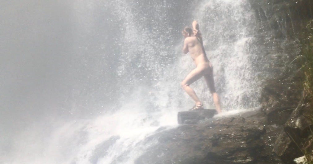 A naked person scrubbing their armpit under a waterfall