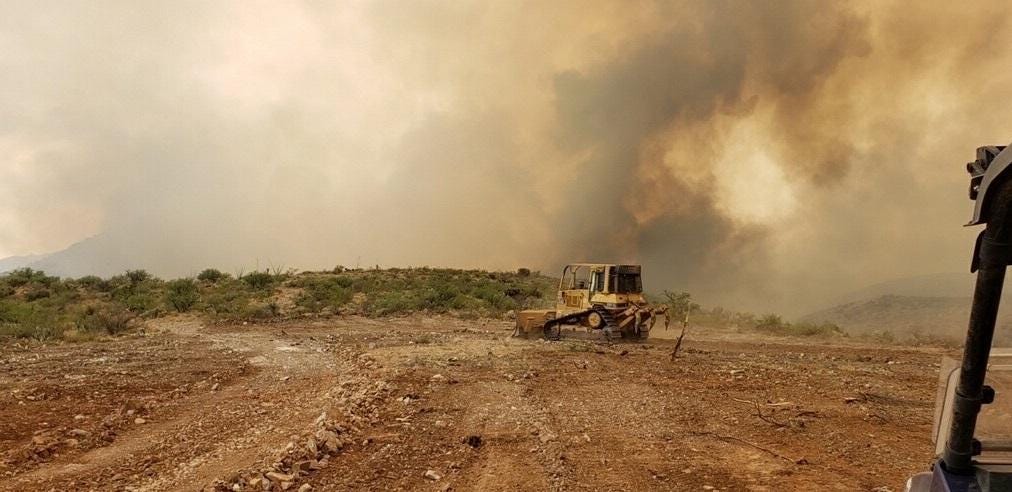 A bulldozer on a stretch of brown dirt, in front of a sky blotted out by smoke clouds.