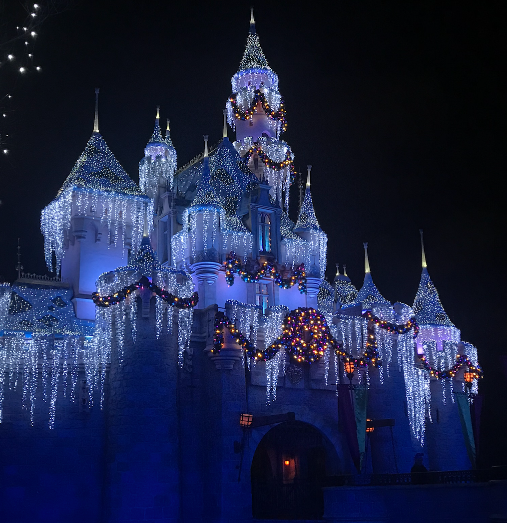 Night shot of Sleeping Beauty Castle at the Disneyland Resort decorated for the holidays with white icicle lights, multi-color lit garland, and a wreath.