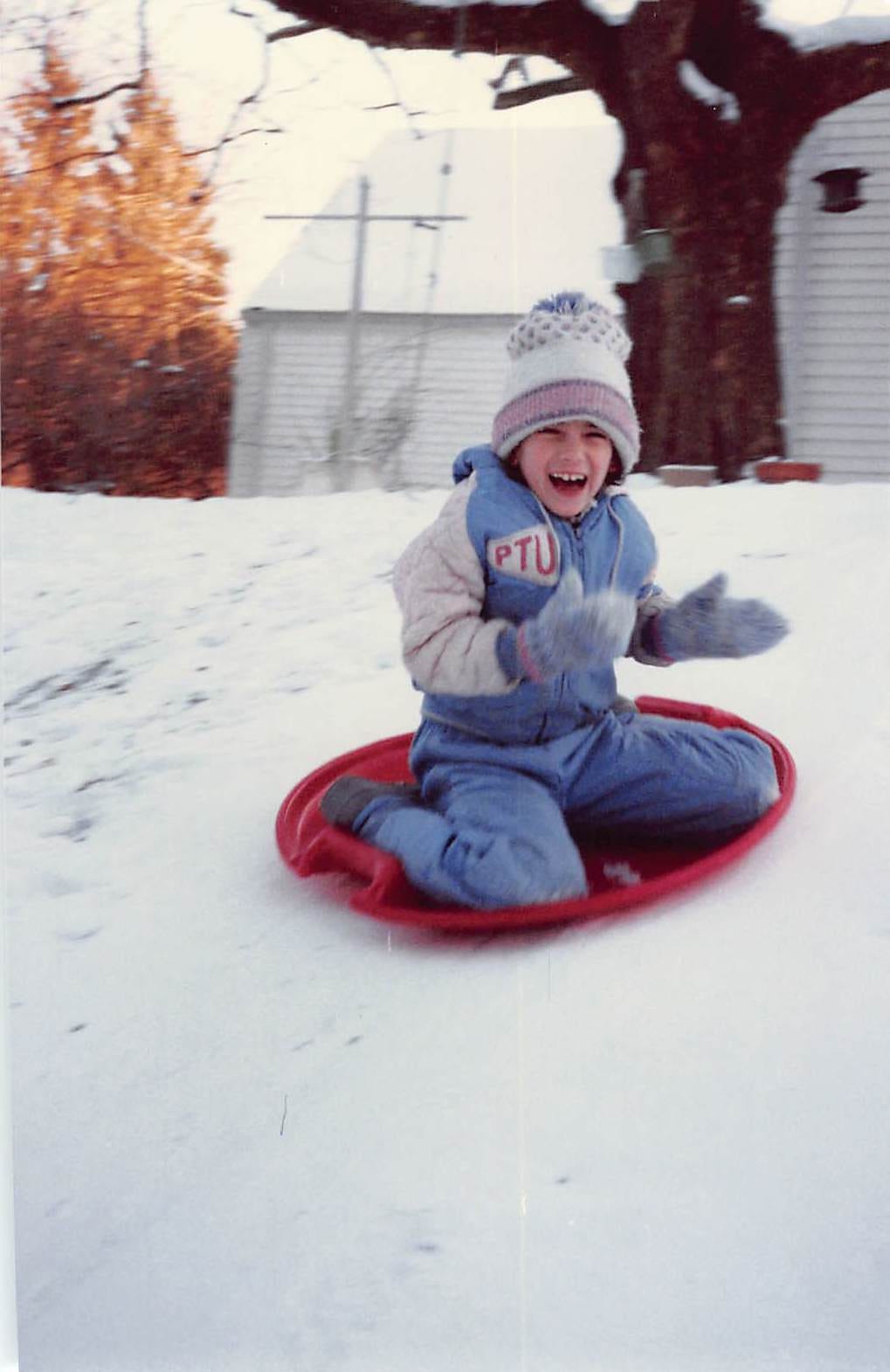 Young child in a snowsuit, sledding down a snowy hill on a red saucer sled, with an expression of utter delight on her face.