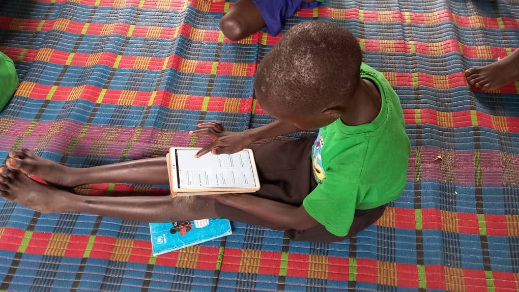 lateral bird’s eye view of a boy sitting on a mat on the floor, engaging with a tablet. A workbook by his side.