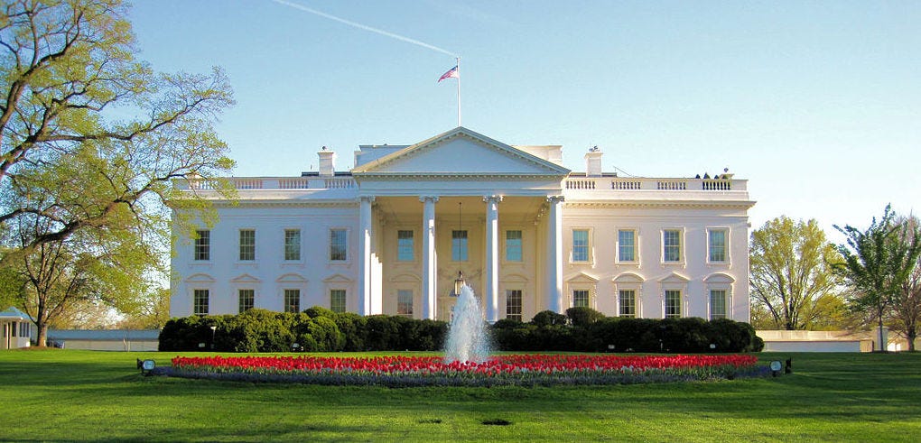 Photo of the White House from the North on a sunny day. There is a fountain surrounded by red flowers in the foreground.