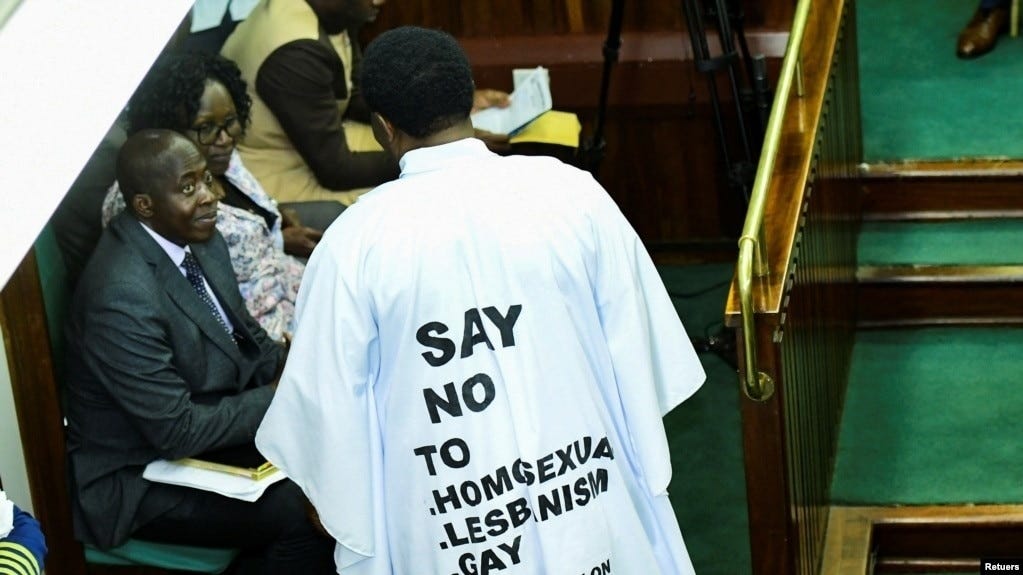 Person in white robe printed Say No to Homosexuality, Lesbianism, Gay. Appears to be in a government chamber or courtroom, talking to two seated people.